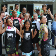 A group of children stand with artist smocks, smiling and making peace signs at the camera.